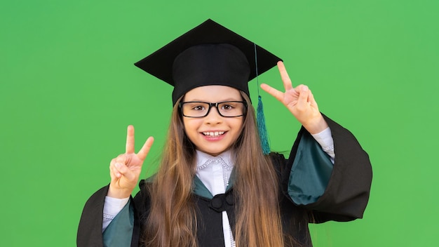 A young schoolgirl in a graduation cap and a ceremonial robe and smiles and shows two fingers on her hands. on an isolated green background.