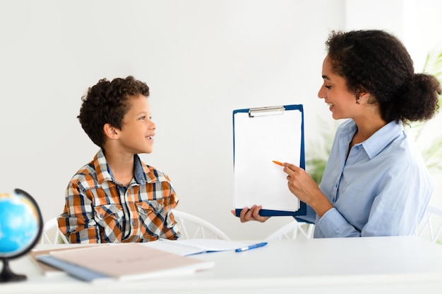 Young schoolteacher woman showing blank paper clipboard to schoolboy indoor