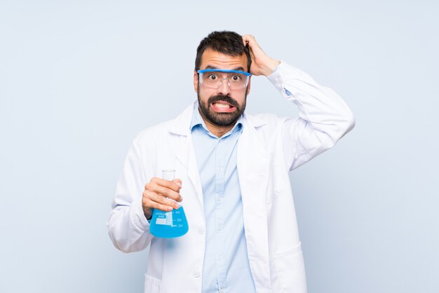 Young scientific holding laboratory flask over isolated wall frustrated and takes hands on head