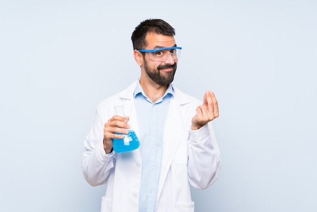 Young scientific holding laboratory flask over isolated wall making money gesture