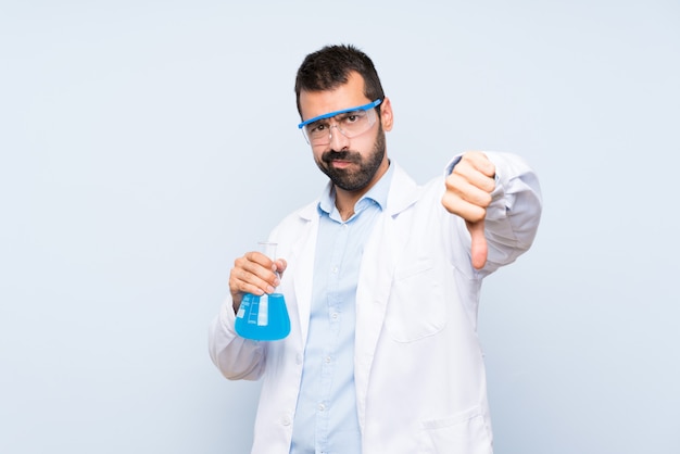 Young scientific holding laboratory flask over isolated wall showing thumb down with negative expression