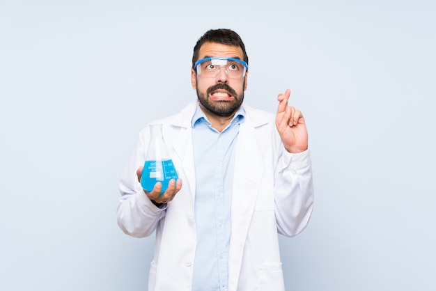 Young scientific holding laboratory flask over isolated wall with fingers crossing and wishing the best