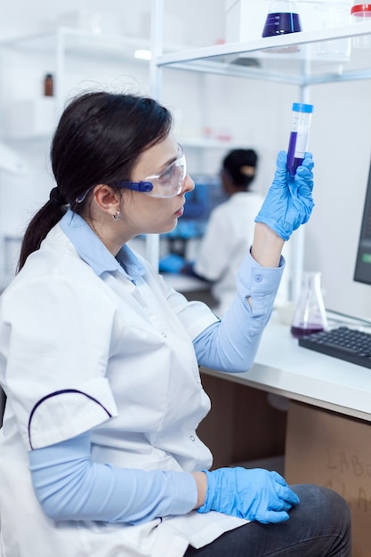 Young scientist analysing scientific sample on plastic test tube with african coworker sitting at her workplace. Chemistry researcher in sterile laboratory using modern technology to test microbiology