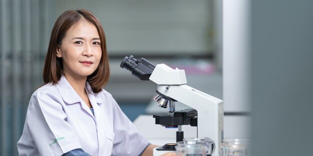 연구와 실험을 하기 위해 실험실에서 현미경을 들여다보고 있는 실험실 코트를 입은 젊은 과학자 여성. 실험실에서 일하는 과학자. 교육 스톡 사진