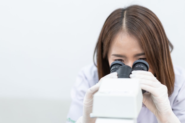 研究と実験を行うために実験室の顕微鏡を通して見ている白衣を着た若い科学者の女性。実験室で働く科学者。教育ストックフォト