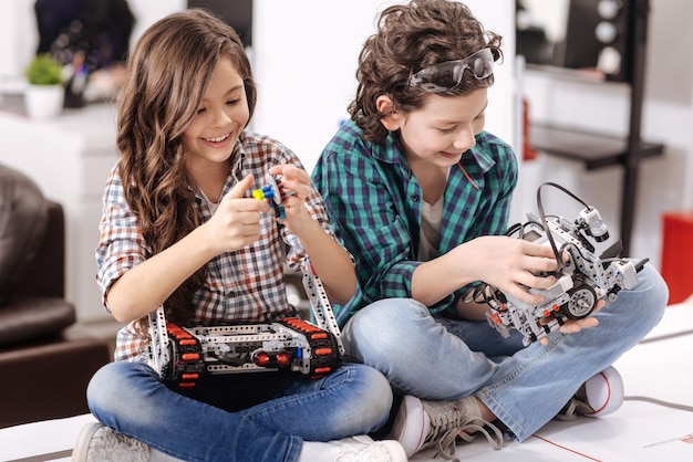 Young scientists cooperating. Smiling cheerful delighted kids sitting at home and testing gadgets and devices while expressing joy