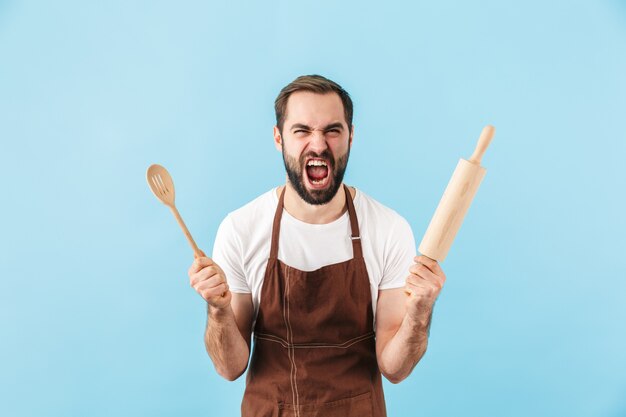 麺棒と木のスプーンを保持している青い壁に隔離された若い悲鳴を上げる感情的な男のシェフ。