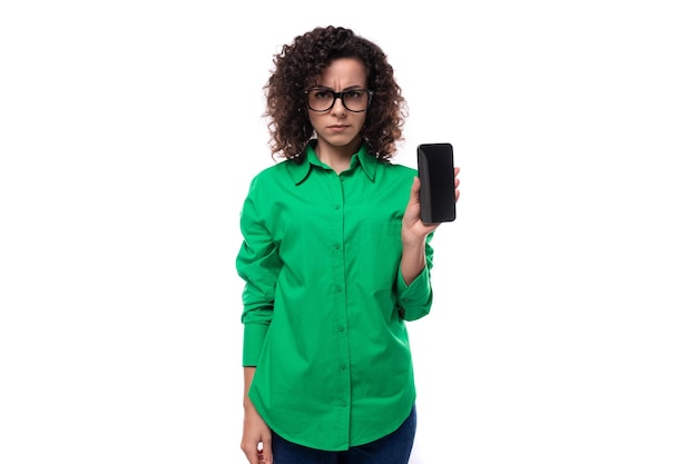 Молодая секретарша брюнетка с кудрявыми волосами, одетая в зеленую рубашку, показывает экран мобильного телефона
