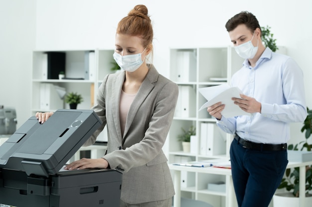 Young secretary in elegant suit and protective mask putting paper in xerox machine to make copies with male colleague waiting on background