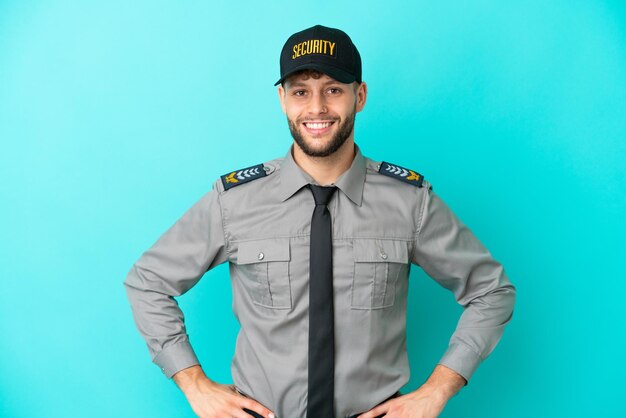 Young security man isolated on blue background posing with arms at hip and smiling