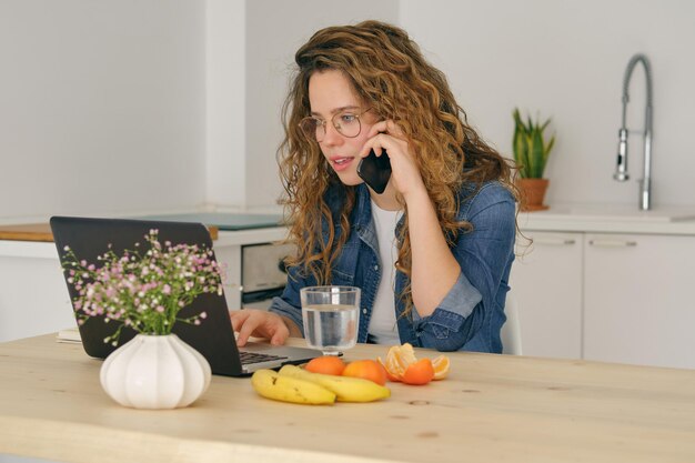 Young self employed woman in casual clothes and eyeglasses sitting at table while speaking on mobile phone and working on laptop at home