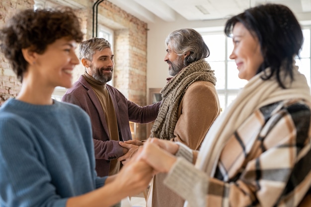 Young and senior men and women in casualwear greeting one another by handshake and looking at each other against home environment