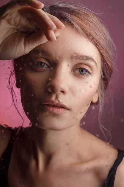 young sensual girl puts her face on her hand and leans on glass on which blurred drops of water flow down. sad concept