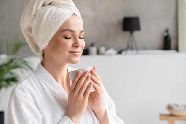 Photo young serene relaxed woman in spa bath towel drinking hot beverage tea coffee after taking shower bath at home beauty treatment hydration concept