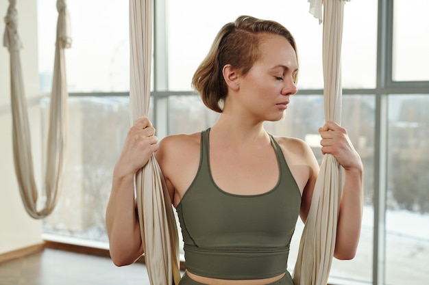 Young serene woman in grey tracksuit keeping her eyes closed\
and holding by fly yoga hammock in gym while exercising in front of\
camera