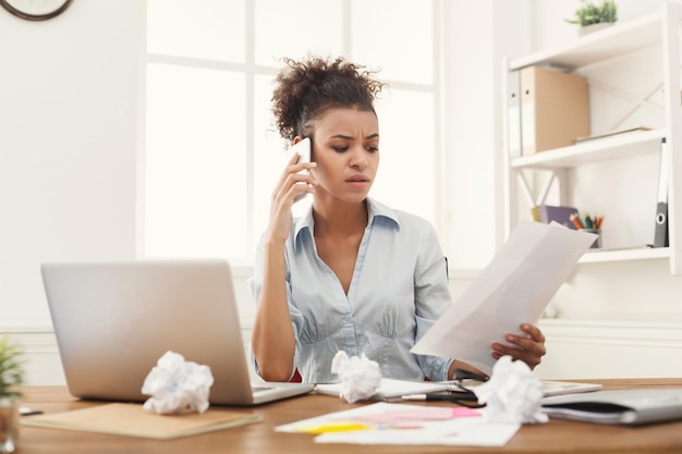 Young serious african-american businesswoman talking by phone with papers, sitting at modern office workplace. Business consulting, copy space