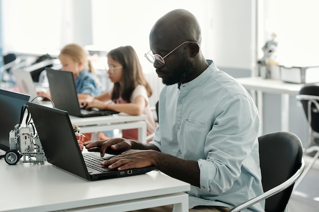 Giovane insegnante di robotica afroamericano serio seduto davanti al laptop