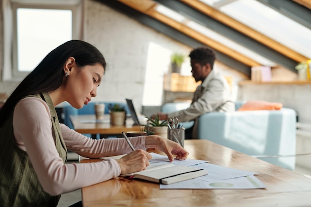 Young serious asian businesswoman with pen making notes in notebook