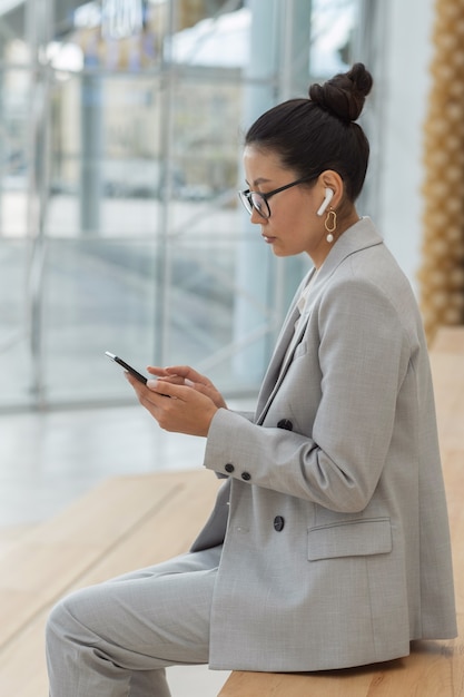 Young serious asian female in suit eyeglasses and earphones using smartphone