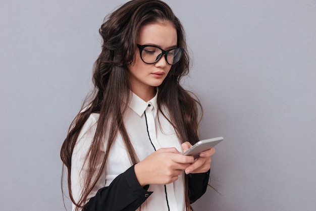 Young serious Asian woman in glasses writing message on phone isolated on gray