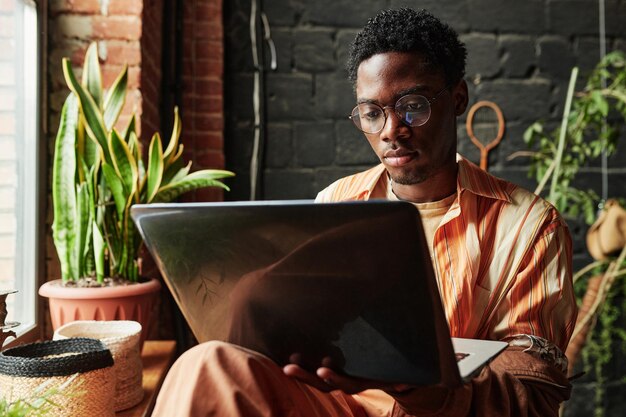 Photo young serious black man in casualwear and eyeglasses using laptop at home