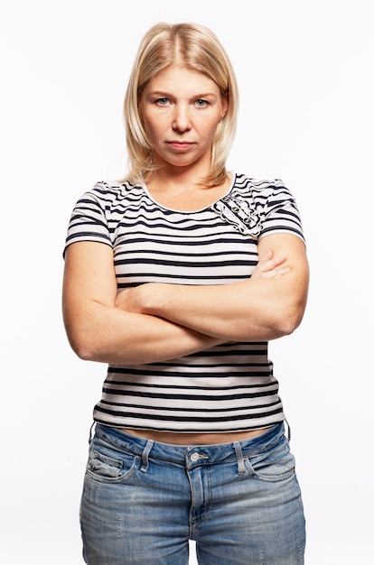 Young serious blonde woman in jeans crossed her arms over her chest. White wall. Vertical.