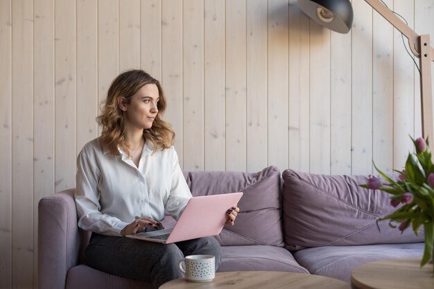 young serious brownhaired woman works at home online sitting on couch she reflects on letter to client looking out window work at home online modern technologies