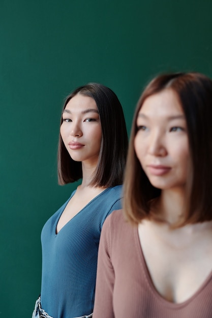 Young serious brunette female of Asian ethnicity with long hair looking at you while standing behind her gorgeous twin sister in studio