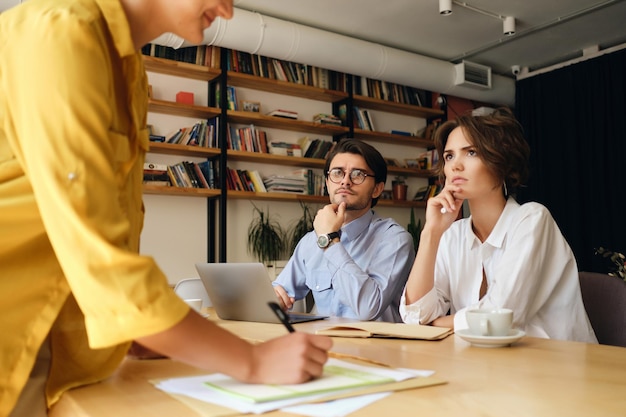 Giovani colleghi di lavoro seri seduti alla scrivania con il laptop che guardano pensierosamente il capo mentre discutono insieme di un nuovo progetto in un ufficio moderno