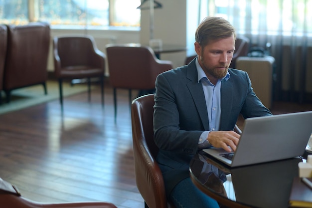 Young serious businessman in formalwear sitting by table in front of laptop