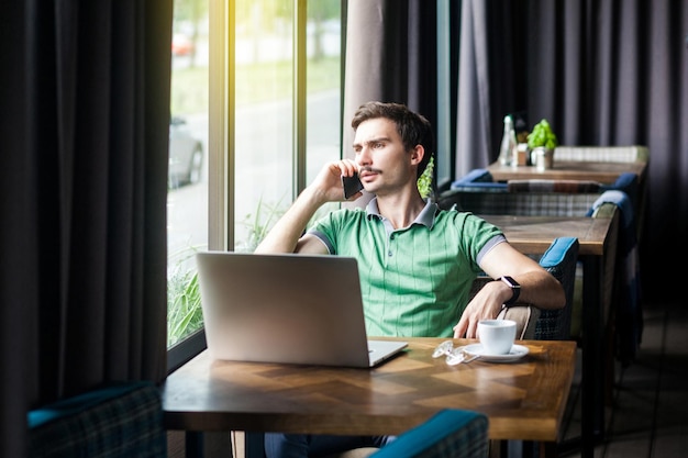 Young serious businessman in green t-shirt sitting at desk with laptop, talking on phone and looking outside. business and freelancing concept. indoor shot near big window at daytime.