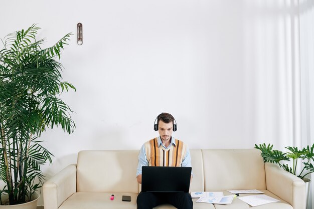 Young serious businessman in headphones sitting on sofa in living room and working on laptop