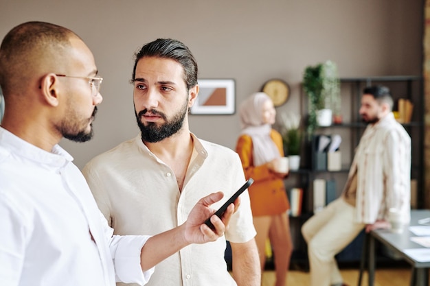 Young serious businessman looking at colleague with smartphone