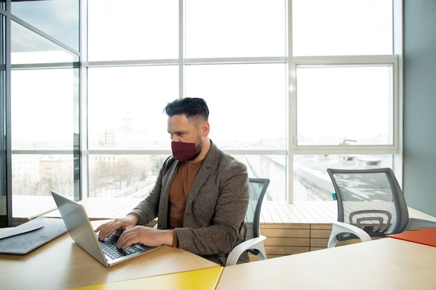 Young serious businessman in protective mask and casualwear sitting in front of laptop by desk against large window and networking