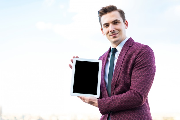 赤いスーツとネクタイとシャツの若い真面目な実業家は空のタブレットで屋根の上に立つ