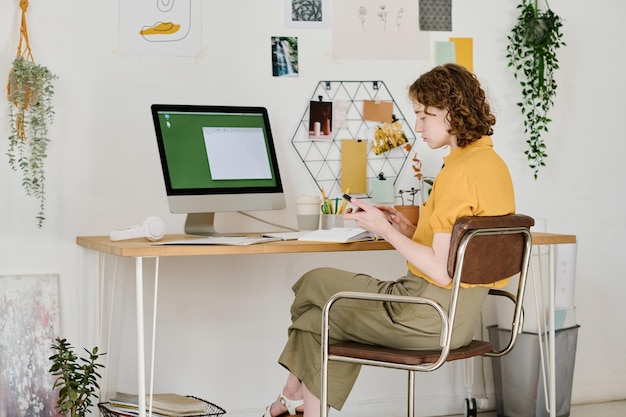 Young serious businesswoman scrolling in mobile phone in front of computer