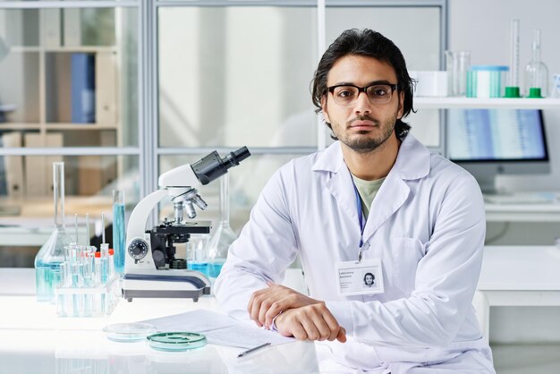 Молодой серьезный клиницист в лабораторном халате и очках сидит на рабочем месте