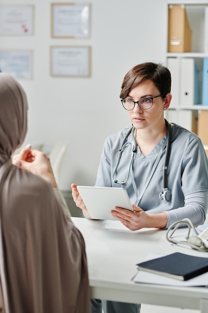 Young serious clinician with tablet consulting muslim female patient in hijab