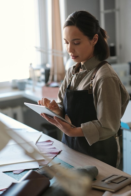 Young serious female artisan in workwear scrolling in tablet by workplace