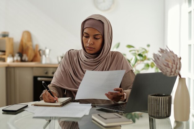 Young serious female auditor in hijab making notes in copybook while sitting by workplace