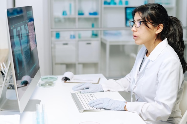 Young serious female researcher or virologist in whitecoat and gloves at work