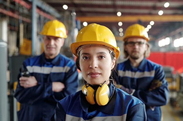 Young serious female worker of large industrial plant or factory in workwear and protective helmet standing against two male colleagues