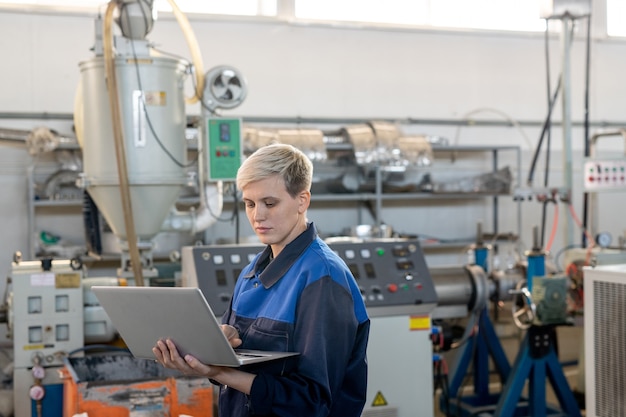 산업 기계에 대한 온라인 기술 데이터를 살펴보는 동안 노트북 디스플레이를 보고 있는 작업복을 입은 젊은 진지한 여성 노동자