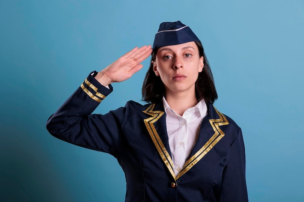 Young serious flight attendant saluting, wearing uniform,\
standing, looking at camera. confident plane stewardess portrait,\
serious air hostess with hand near head front view, studio medium\
shot