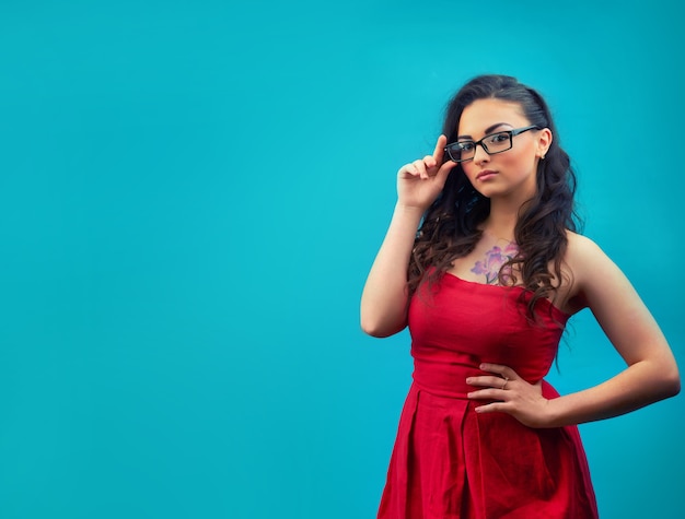 Young serious girl with makeup, wearing glasses and a red dress on a blue wall