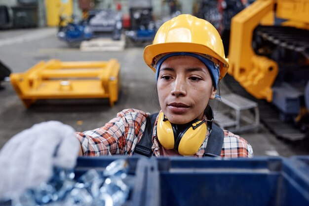 Young serious hispanic female factory worker in gloves hardhat and overalls