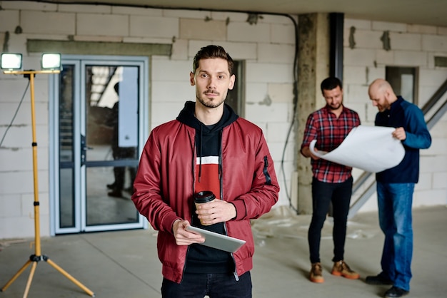 Young serious house designer with drink and tablet standing in front of camera while his colleagues discussing sketch on blueprint