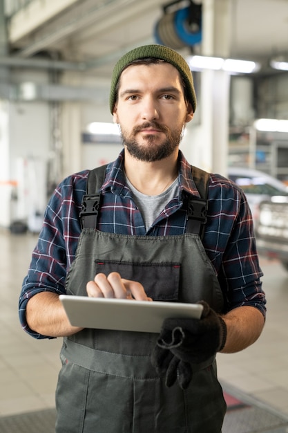 Foto giovane lavoratore maschio serio del servizio auto che utilizza tablet digitale mentre risponde alle domande online dei clienti nell'ambiente di lavoro