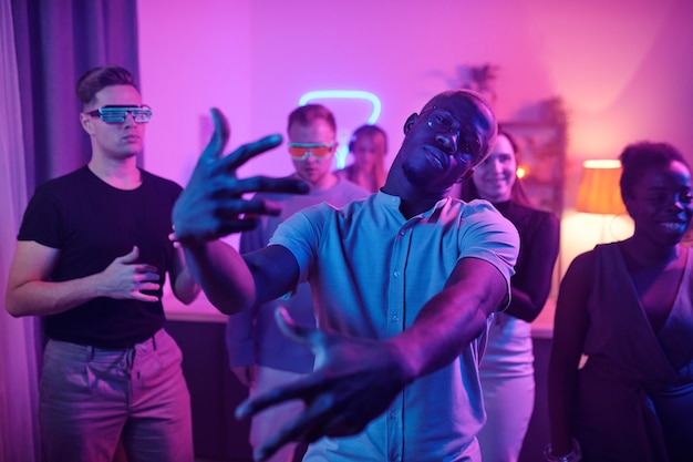 Young serious man of African ethnicity in casualwear and eyeglasses looking at you while standing in front of his friends at home party