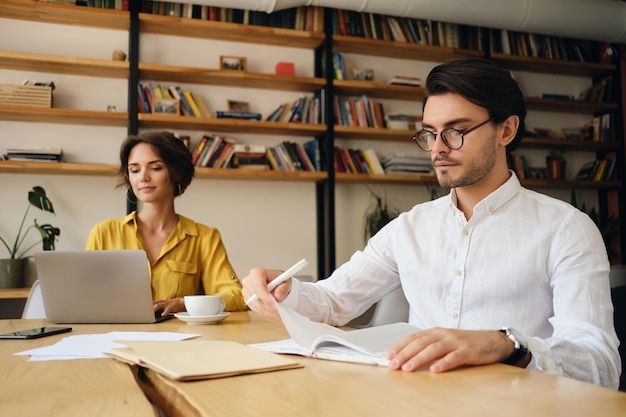 Giovane uomo serio con gli occhiali seduto al tavolo che lavora premurosamente in un ufficio moderno con il collega sullo sfondo